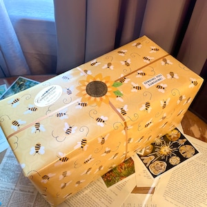 May include: A yellow gift box with a sunflower and bees pattern. The box is wrapped with a brown ribbon and has a sticker that says "Connoisseur Gifts" and "Sending You Sunshine!"