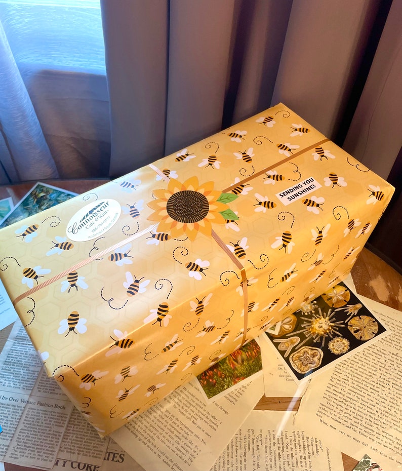 May include: A yellow gift box with a sunflower and bees pattern. The box is wrapped with a brown ribbon and has a sticker that says "Connoisseur Gifts" and "Sending You Sunshine!"