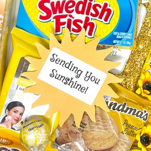 May include: A yellow paper sun cutout with the text "Sending You Sunshine!" on a white background. The sun is surrounded by various yellow and gold items, including sweets, snacks, and a bar of soap.