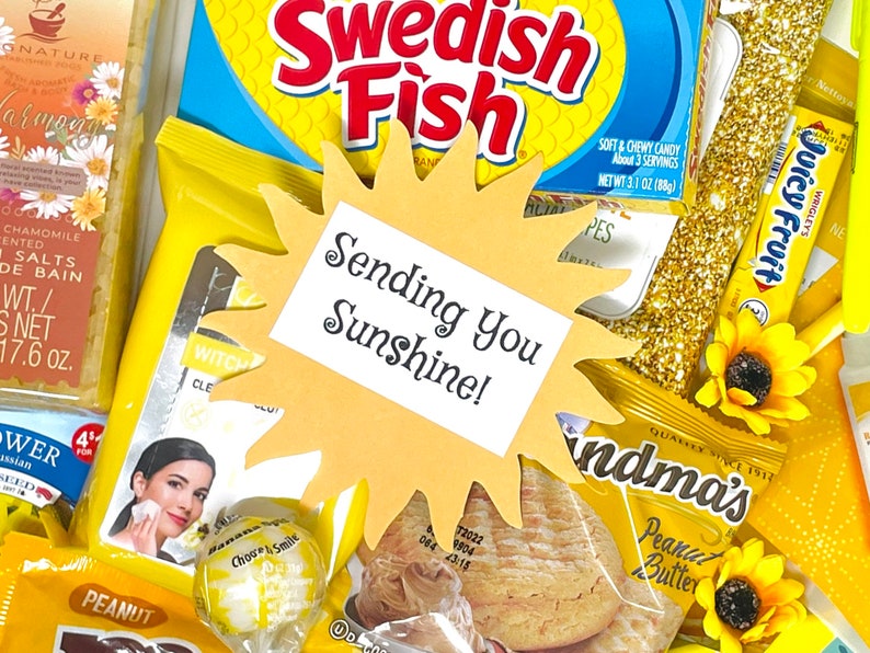 May include: A yellow paper sun cutout with the text "Sending You Sunshine!" on a white background. The sun is surrounded by various yellow and gold items, including sweets, snacks, and a bar of soap.