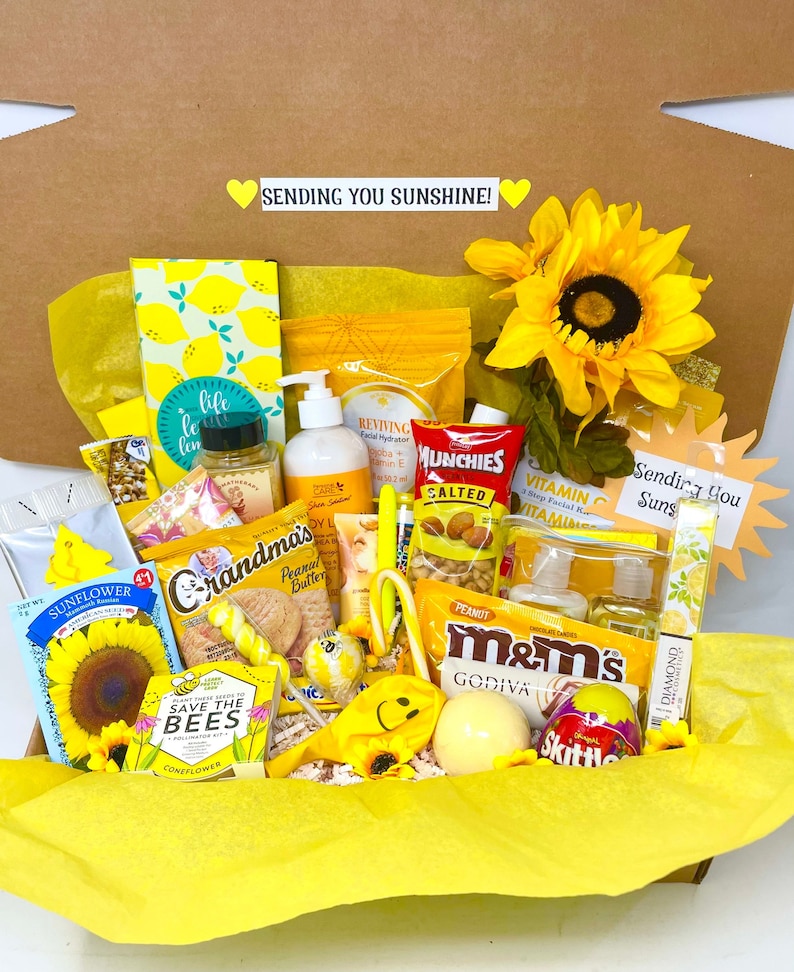 May include: A yellow gift box filled with various items, including snacks, bath products, and a sunflower. The box is decorated with yellow and white tissue paper and a yellow ribbon. The text "Sending You Sunshine!" is printed on the box.