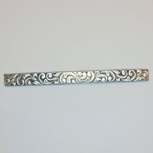 May include: A silver bar with a decorative, engraved floral pattern.