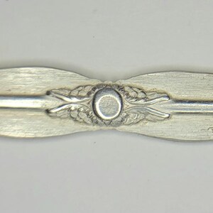 May include: Close-up of a silver-toned metal spoon handle with a decorative design featuring a circle in the center and leaf-like patterns on either side.