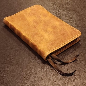 May include: A tan leather-bound book with a textured cover and dark brown ribbon bookmarks. The book is closed and lies on a dark brown leather surface. The edges of the book are a lighter tan color.