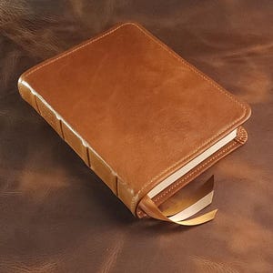 May include: A closed, rectangular, brown leather-bound book with a gold ribbon bookmark. The book has a stitched spine and a smooth, polished cover. The book is resting on a brown leather surface.