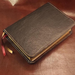 May include: A closed, dark brown leather-bound book with gold-colored stitching around the edges. Two colored ribbon bookmarks are visible. The book is on a brown leather surface.