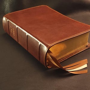 May include: A brown leather-bound Holy Bible with gold-gilded pages and three attached ribbon bookmarks. The book has gold lettering on the spine and is resting on a dark surface.