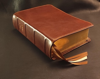 May include: A brown leather-bound Holy Bible with gold-gilded pages and three attached ribbon bookmarks. The book has gold lettering on the spine and is resting on a dark surface.