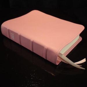 May include: A pink leather-bound book with a ribbon bookmark. The book has a smooth cover and a spine with embossed details. The pages are white, and the bookmark is a light pink color. The book is set against a black background.