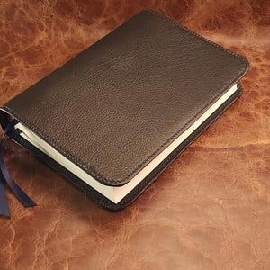 May include: A dark brown leather-bound journal with a textured surface. The journal has a blue ribbon bookmark and white-edged pages. The cover is closed, and the journal is resting on a brown leather surface.