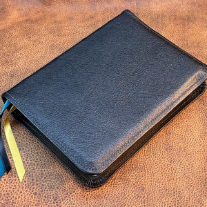 May include: A closed, black leather-bound book with a textured surface. Three colored ribbon bookmarks, blue, black, and yellow, extend from the top. The book rests on a brown, textured surface.