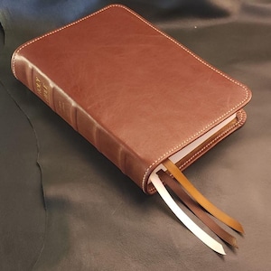 May include: A brown leather-bound book with gold lettering on the spine. The book has three colored ribbon bookmarks and white stitching around the edges. The book is resting on a dark surface.