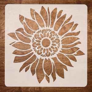 May include: A white stencil of a sunflower with a detailed center and petals. The stencil is on a brown wooden background.