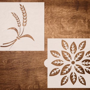 May include: Two white stencils, one with a wheat design and the other with a flower design. The stencils are on a brown wooden background.