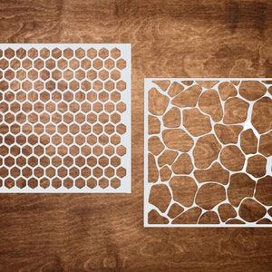 May include: Two white stencils with different patterns. One features a honeycomb design, and the other has an irregular, organic pattern. The stencils are displayed on a dark brown wooden surface.