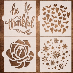 May include: Four white stencils with cut-out designs. One stencil reads "be thankful" with wheat illustrations. Other stencils feature hearts, a rose, and snowflakes with stars. The stencils are on a brown wooden surface.