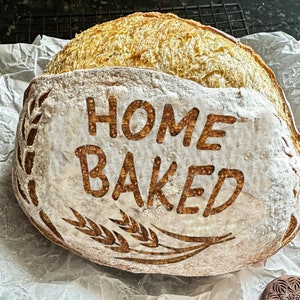 May include: A loaf of bread with the words "Home Baked" written on it in a brown font. The bread is dusted with flour and has a rustic appearance.