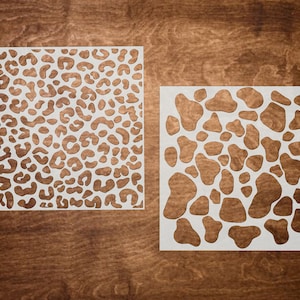 May include: Two white stencils on a brown wooden background. The stencil on the left has a leopard print pattern. The stencil on the right has a pattern of irregular, rounded shapes.
