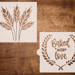 May include: Two white stencils for baking. One stencil features a cluster of wheat stalks. The other stencil features a wreath with the text "Baked with love".