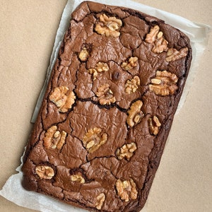 Puede incluir: Un brownie reci&eacute;n horneado, rectangular, con una superficie agrietada y texturizada. El postre de chocolate negro est&aacute; cubierto con varias mitades de nueces. El brownie descansa sobre un cuadrado de papel de pergamino blanco.