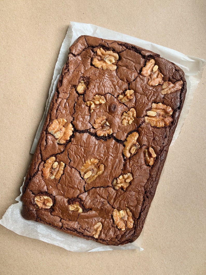 Puede incluir: Un brownie reci&eacute;n horneado, rectangular, con una superficie agrietada y texturizada. El postre de chocolate negro est&aacute; cubierto con varias mitades de nueces. El brownie descansa sobre un cuadrado de papel de pergamino blanco.