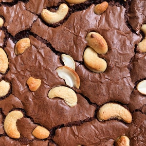 Puede incluir: Primer plano de un brownie reci&eacute;n horneado con una superficie agrietada. El brownie es de un color marr&oacute;n oscuro intenso y est&aacute; generosamente cubierto con anacardos enteros. Las nueces son de color marr&oacute;n claro y a&ntilde;aden textura al postre.