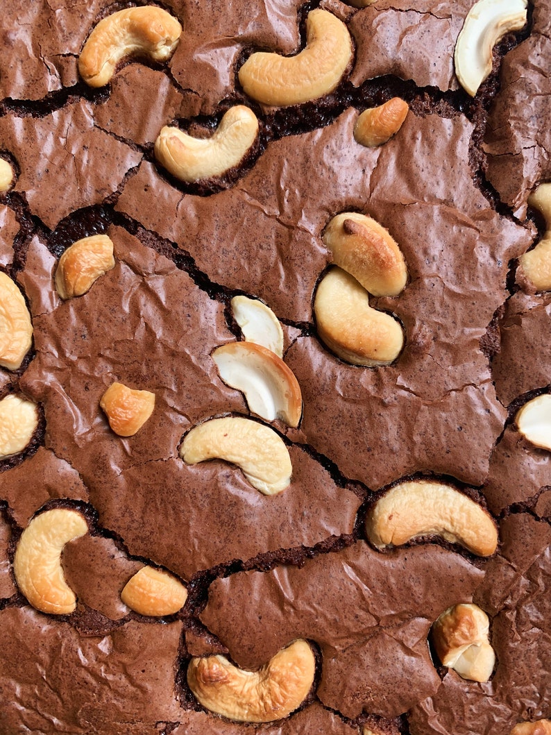 Puede incluir: Primer plano de un brownie reci&eacute;n horneado con una superficie agrietada. El brownie es de un color marr&oacute;n oscuro intenso y est&aacute; generosamente cubierto con anacardos enteros. Las nueces son de color marr&oacute;n claro y a&ntilde;aden textura al postre.