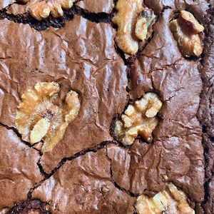 Puede incluir: Primer plano de un brownie reci&eacute;n horneado con una superficie agrietada y texturizada. El brownie es de un rico color marr&oacute;n oscuro y est&aacute; cubierto con varios trozos de nueces doradas. La imagen destaca la calidad casera del postre.