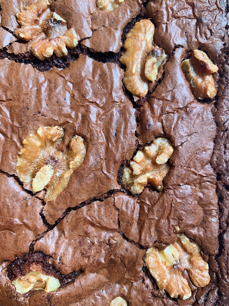 Puede incluir: Primer plano de un brownie reci&eacute;n horneado con una superficie agrietada y texturizada. El brownie es de un rico color marr&oacute;n oscuro y est&aacute; cubierto con varios trozos de nueces doradas. La imagen destaca la calidad casera del postre.