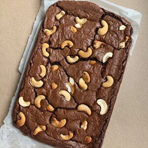 Puede incluir: Un brownie de chocolate rectangular cubierto con anacardos, sobre papel de pergamino blanco. El brownie es de color marr&oacute;n oscuro con una superficie agrietada, y los anacardos dorados est&aacute;n esparcidos por encima.