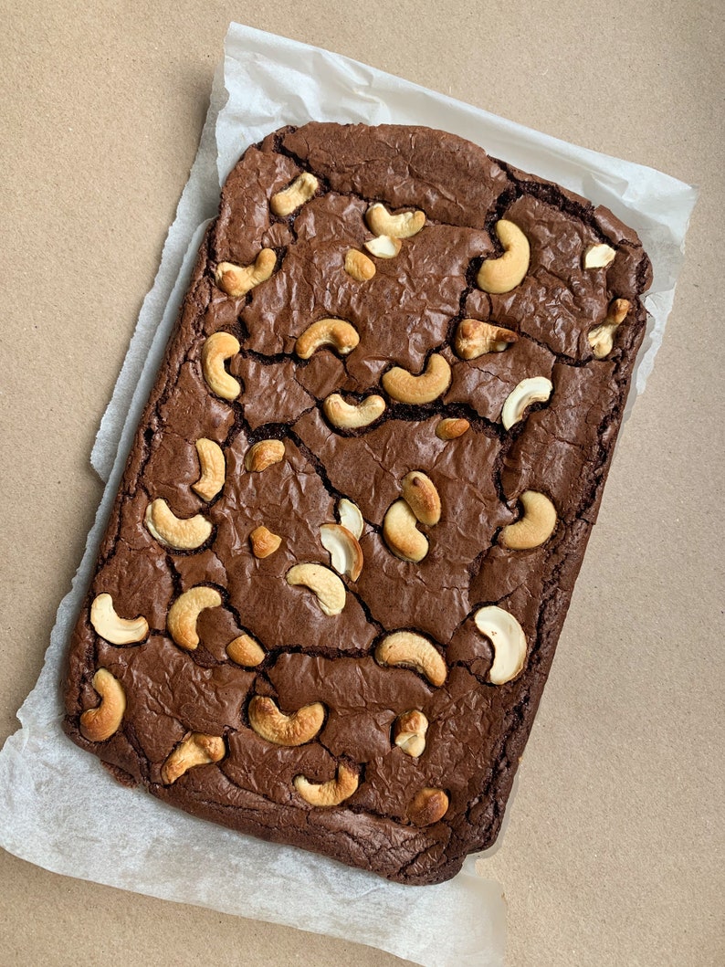 Puede incluir: Un brownie de chocolate rectangular cubierto con anacardos, sobre papel de pergamino blanco. El brownie es de color marr&oacute;n oscuro con una superficie agrietada, y los anacardos dorados est&aacute;n esparcidos por encima.