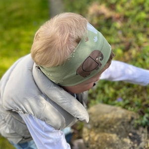 May include: A light green headband featuring a repeating pattern of brown elephants and white shapes. The headband is worn by a person wearing a white long-sleeve shirt and a gray vest. The background includes green grass and foliage.