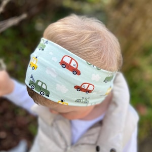 May include: A light blue headband with a repeating pattern of cartoon cars in red, green, yellow, and blue. The cars are interspersed with white clouds. The headband is worn by a child.