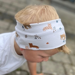 May include: White headband with a repeating pattern of brown and tan animal illustrations and green leaf accents. The headband is made of a soft fabric and is designed to fit comfortably around the head.