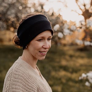 May include: A black ribbed headband worn by a person with light skin and brown hair. The person is wearing a beige sweater and is smiling. The background is a blurred outdoor scene with trees and sunlight.