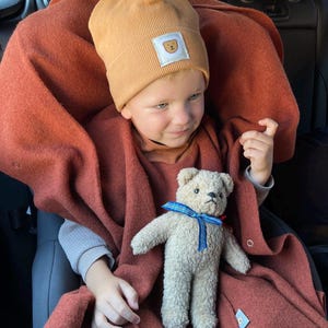 May include: A child wearing a mustard yellow beanie with a teddy bear patch and a rust-colored blanket. A stuffed teddy bear with a blue ribbon sits in the child's lap. The child is seated in a car.