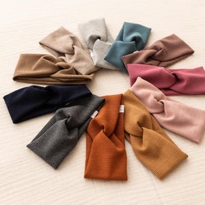 May include: A collection of ten ribbed knit headbands in various colors, including tan, gray, blue, brown, and pink. The headbands are arranged in a circular pattern on a light-colored surface, showcasing their twisted knot design.