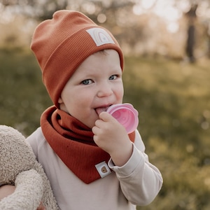 May include: A young child wearing a rust-colored beanie and matching scarf, holding a plush toy and chewing on a pink teether.
