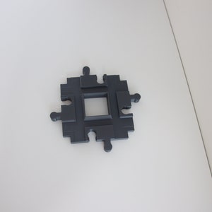 Könnte beinhalten: Ein quadratisches Spielzeug-Eisenbahnstück aus schwarzem Kunststoff mit vier Verbindungspunkten an jeder Seite.