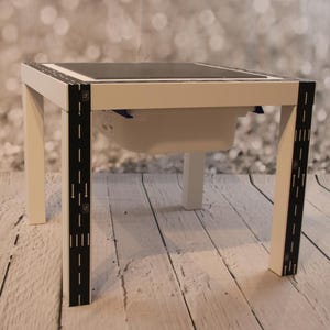 Peut inclure: Table blanche avec un plateau de base en Lego noir et gris. La table a un bac de rangement blanc en dessous et du ruban adhésif noir sur les pieds avec des marques qui ressemblent à une route.