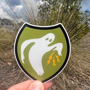 May include: A sticker featuring a white ghost design with orange lightning bolts emanating from its mouth. The ghost is set against a green background within a black-bordered shield shape. The sticker is held up against a backdrop of tall grass and a cloudy sky.