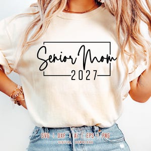 May include: A cream-colored t-shirt featuring a black graphic design with the text Senior Mom 2027 enclosed in a rectangular frame. The shirt has short sleeves and a crew neckline. The text SVG | DXF | AI | EPS | PNG DIGITAL DOWNLOAD is visible.