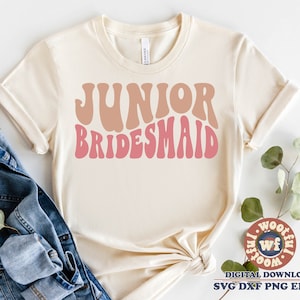 Puede incluir: Una camiseta blanca con el texto "Junior Bridesmaid" en rosa y marrón en fuente retro.