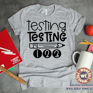 Puede incluir: Una camiseta gris con las palabras "testing TESTING" en negro, un dibujo de lápiz y los números "1 2 3". La imagen incluye útiles escolares como un cuaderno rojo, lápices y una manzana, sobre un fondo gris.