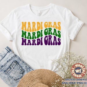 May include: White t-shirt with the words "Mardi Gras" repeated three times in a retro font. The words are in yellow, green, and purple.
