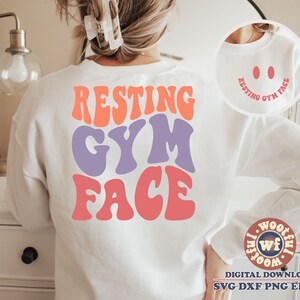 May include: White sweatshirt featuring the text "RESTING GYM FACE" in colourful, retro-style lettering. The text is in a large font and is centred on the sweatshirt. The sweatshirt is being worn by a person, but the person's face is not visible.