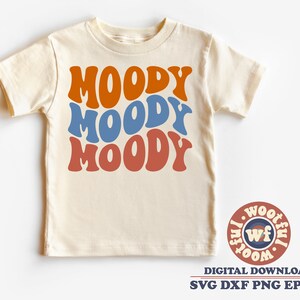 May include: A beige t-shirt with the word "MOODY" written three times in a retro font, with each word in a different color: orange, blue, and red. The shirt is on a white background. The image also includes a logo with the text "WOOTIFUL" in a circle.