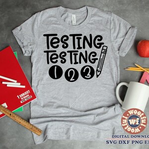 Puede incluir: Camiseta gris con el texto "Testing Testing 123" en negro. El texto está en una fuente de letra manuscrita divertida. Un lápiz blanco y negro está dibujado al lado del texto.
