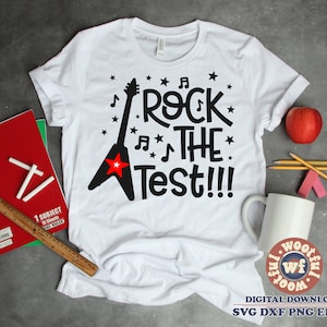 May include: White t-shirt with the text "ROCK THE TEST!!!" in black, featuring a black guitar with a red star. Musical notes and stars surround the text. A red apple, pencils, and a mug are next to the shirt.