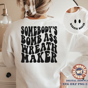 May include: White sweatshirt with black text that reads "Somebody's Bomb Ass Wreath Maker" in a retro font. The text is centered on the back of the sweatshirt.